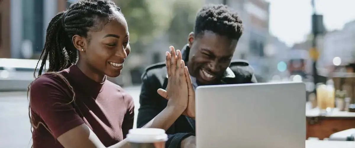 Two students giving high five while working on a laptop outside and drinking coffee