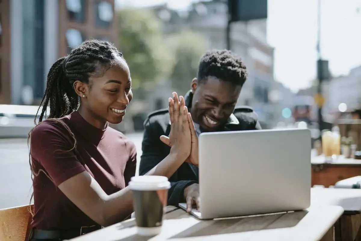 Two students giving high five while working on a laptop outside and drinking coffee